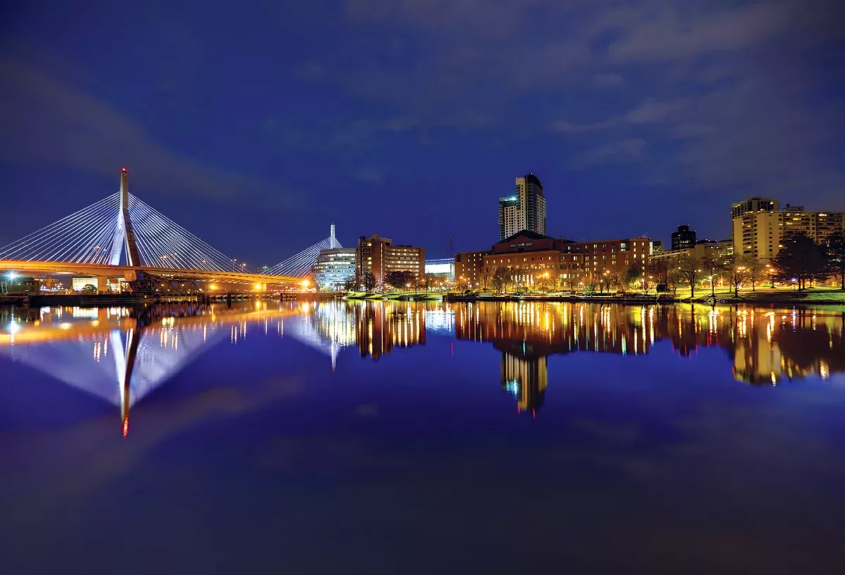 Nighttime city skyline with illuminated buildings reflected on calm water.