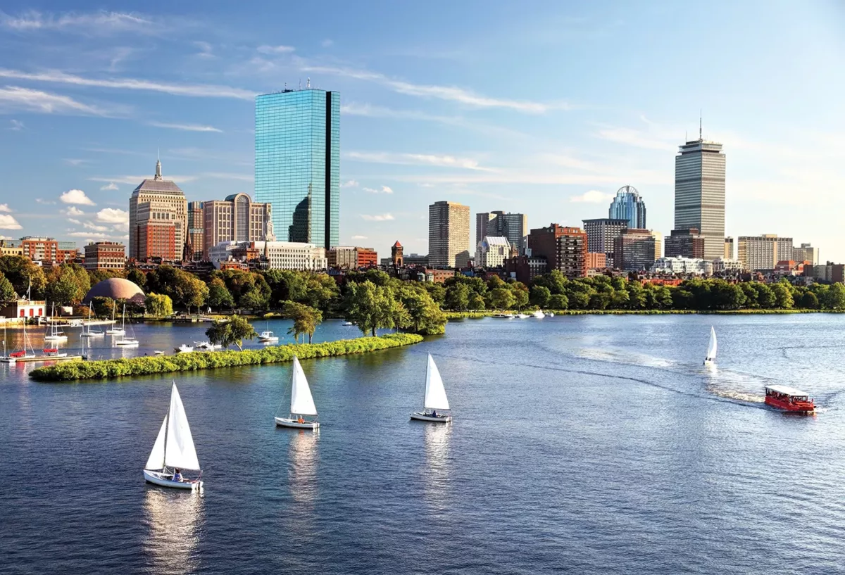 City skyline reflected on river with sailboats and trees along the waterfront.